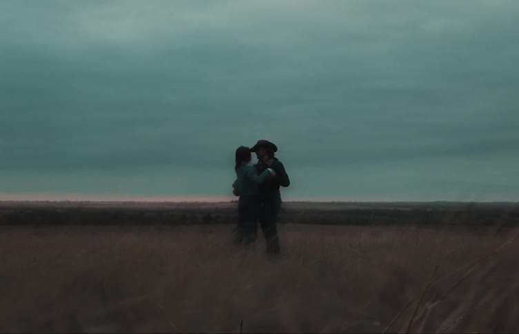 Man wearing cowboy hat pulls woman back into his arms as they stand in field under an overcast sky