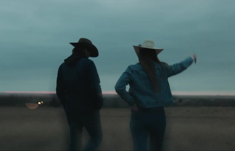 Man and woman wearing cowboy hats talk while walking across field just after dusk