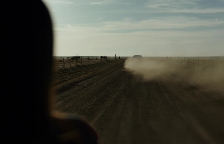 A woman watches as a pickup truck drives away down a dirt road