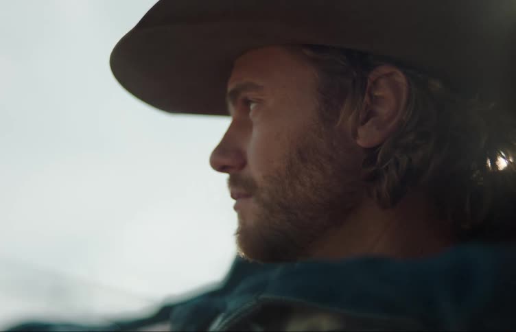 A man standing outside wearing a cowboy hat looking forward contemplatively as the sun shines on his back
