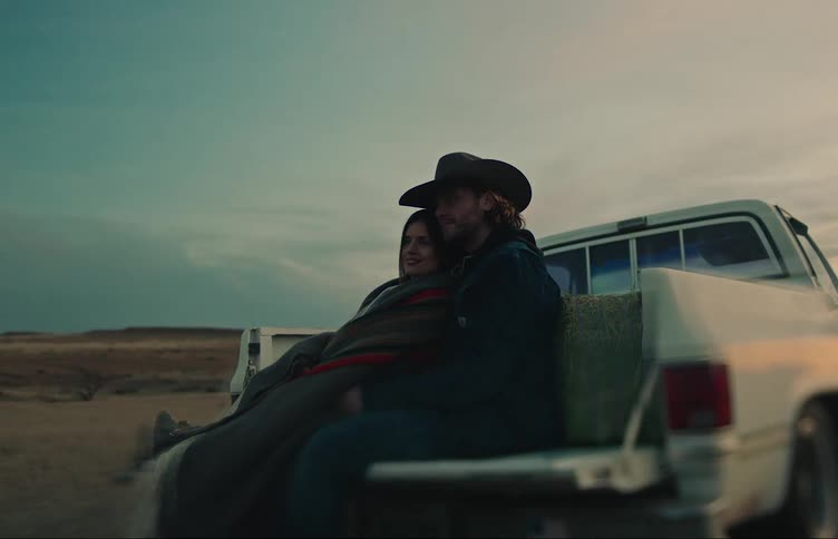 A man and woman sitting on the back of a truck together leaning up against a bail of hay looking out into the distance