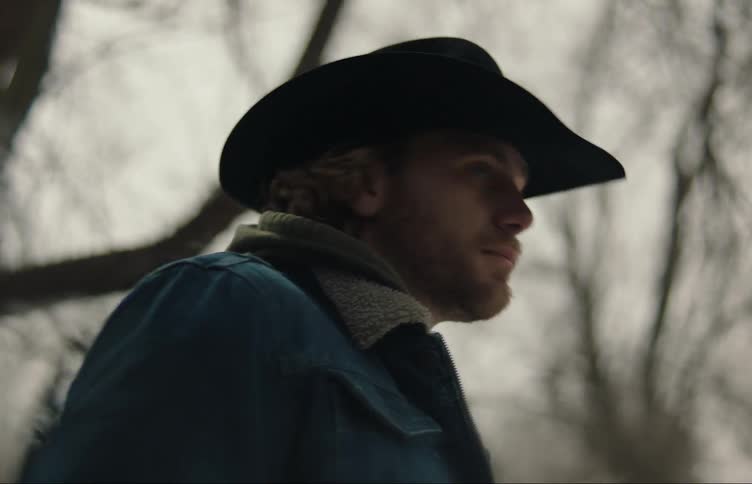 A cowboy looks back and forth while standing in the woods