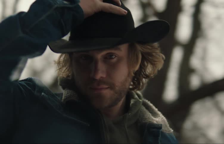 A man standing outside posing for the camera while he puts his cowboy hat on with trees behind him