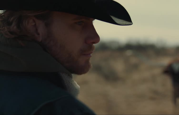 Close up of the face of a cowboy standing next to long horn bulls in the countryside
