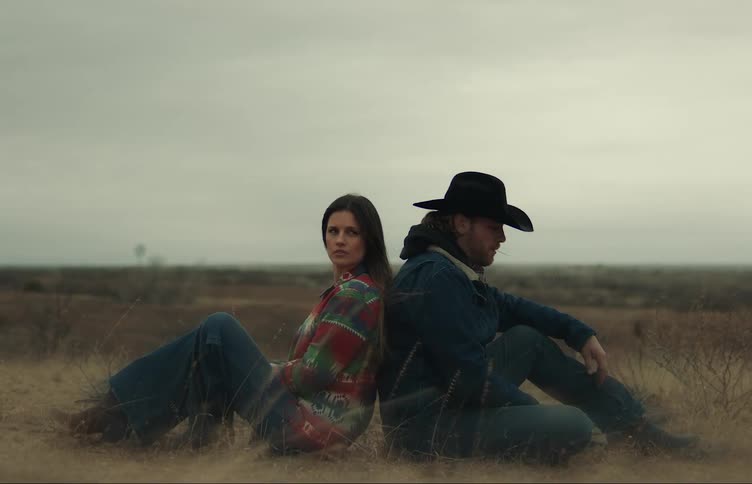 A woman sits back to back with a man wearing a cowboy hat on the empty grass plains