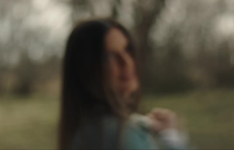 A woman is standing near trees moving her long dark hair with her hand and looking at the camera