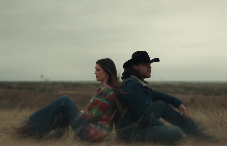 A man and woman sit backtoback in a grassy field in the countryside