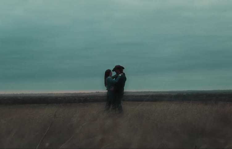 A man wearing a cowboy hat is standing in a rural field with his girlfriend as they have their arms around each other and talk face to face