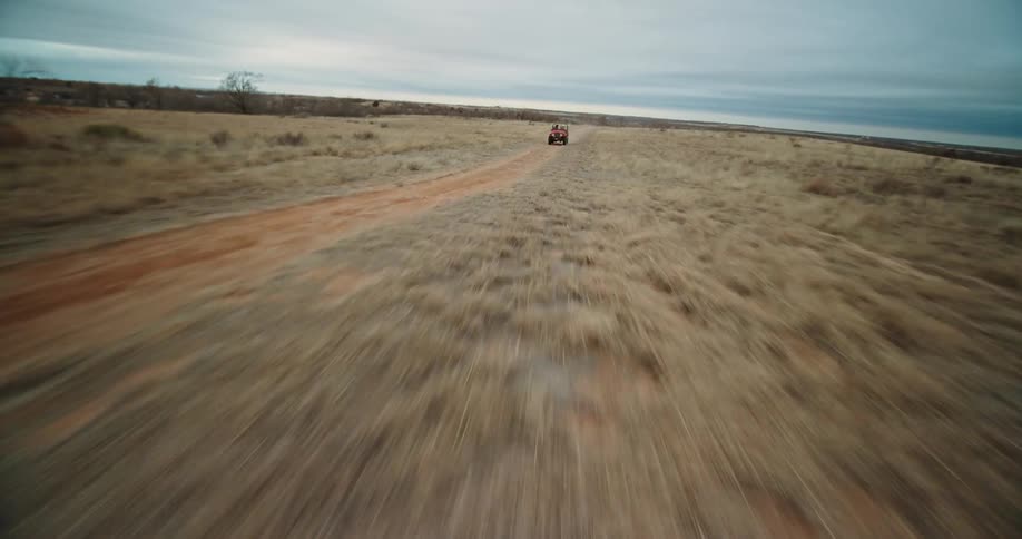 A drone view of a red jeep driving down a dirt road in the desert with two people in the jeep