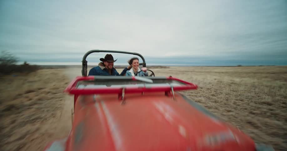 A woman is driving a red jeep on a dirt road near bare trees as a man wearing a cowboy hat sits next to her