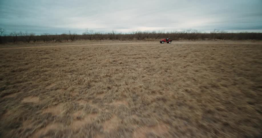 People drive along dirt road between field and leafless trees on cloudy day