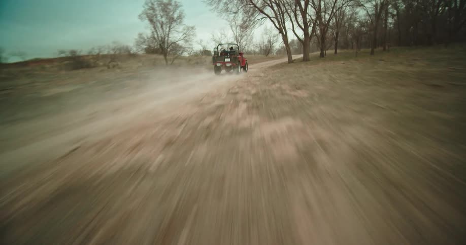 Two people are riding in a red jeep moving fast on a dirt road with bare trees in the woods
