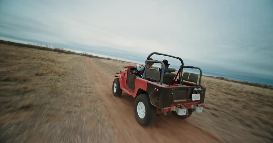 A woman is driving a red jeep on a rural dirt road as a man wearing a cowboy hat sits next to her