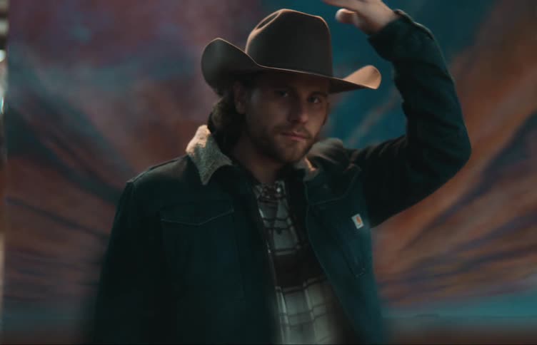 A determined cowboy puts his hat on in front of a painting of a cloudy dawn sky