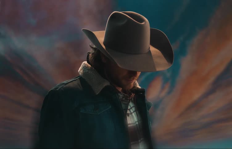 A man wearing a cowboy hat and a jacket is standing in front of a mural
