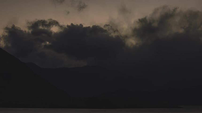 Dark storm clouds move over mountains along the coast as the sunlight fades away