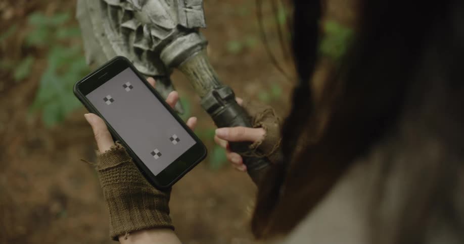A woman wearing fingerless gloves holds a toy ax and a cell phone and presses a symbol on her cell phone screen