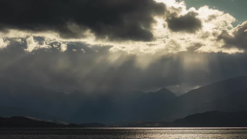 The sun shines through dark clouds as they float over mountains and pour rain into the lake