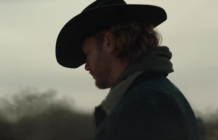 The side profile of a man wearing a cowboy hat walking outdoors and looking toward trees
