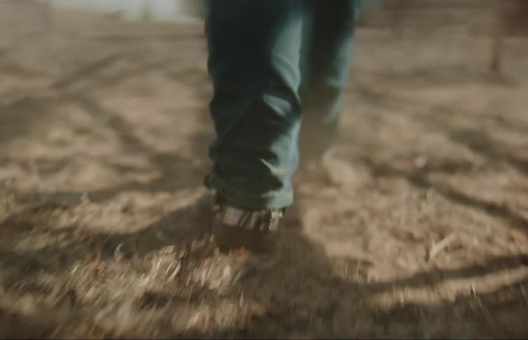 Person wearing boots kicks up dust while walking across dirt toward water