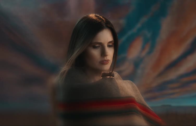 A pensive woman wrapped in a blanket posing in front of a painting of a cloudy dawn sky