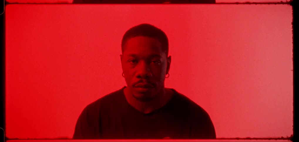 A black man stares intently under red light with his back to a red painting