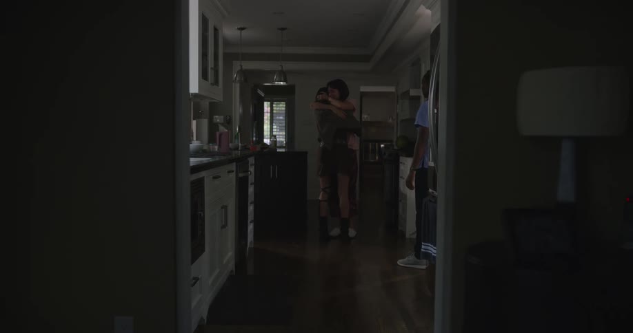 A girl is standing in a room hugging her boyfriend and another person is standing in the kitchen watching them
