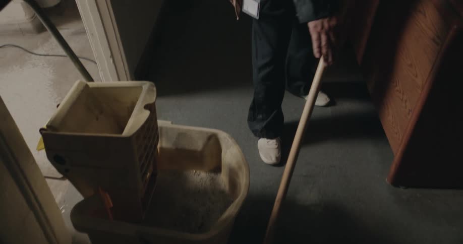 A janitor is standing in a room near a mop bucket as he mops the floor