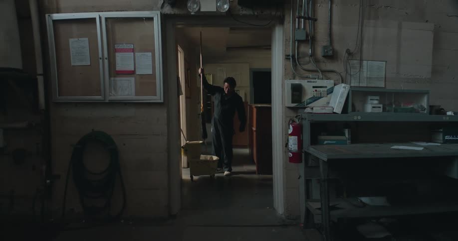 A man is standing in a hallway near a counter mopping the floor and wringing the mop out in the mop bucket