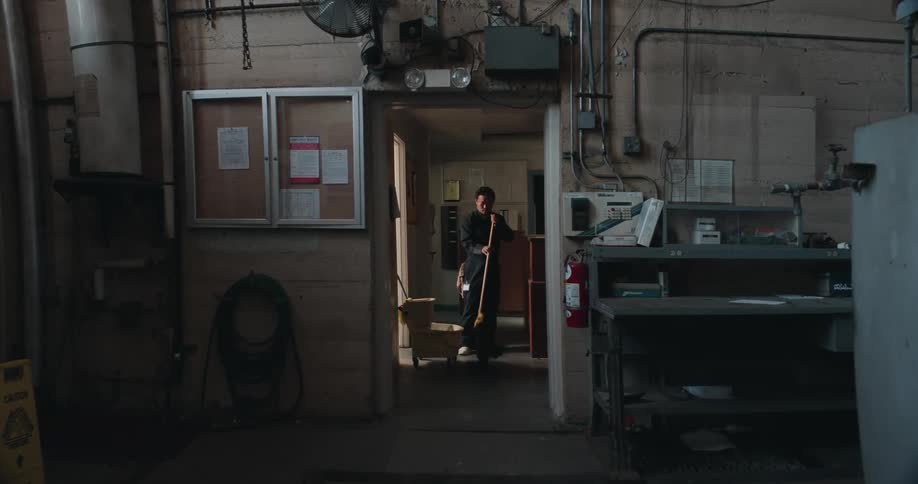 A man is standing in a hallway of a facility near a counter as he mops the floor and pauses to look up