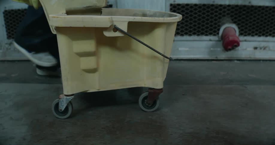 A janitors feet are seen as they walk pushing a mop bucket down the hallway