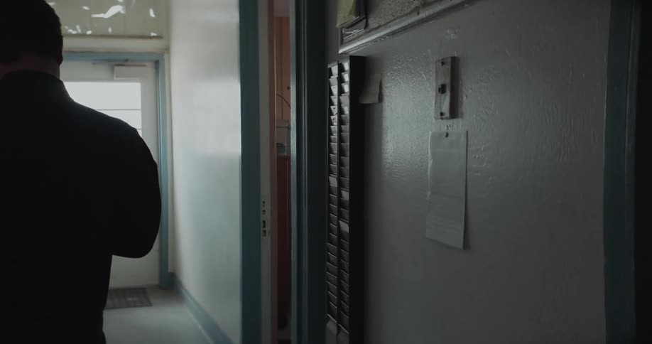 A man is standing in a hallway and takes his time card from the wall and clocks in and puts his card back as he walks