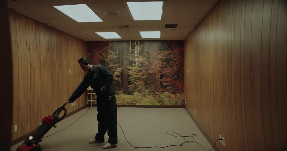 A man is wearing headphones as he vacuums a room with wood paneling and a forest picture on the wall