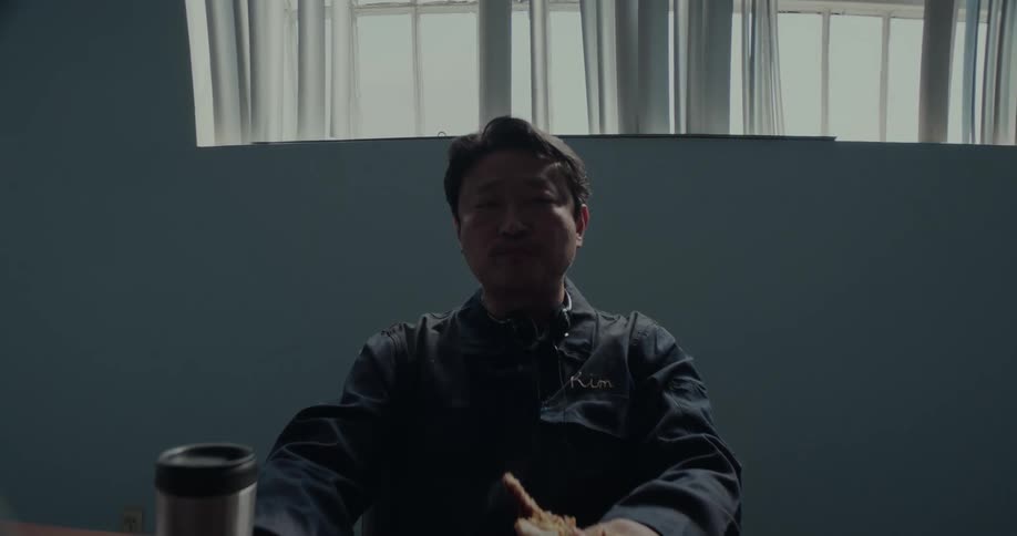 A man is sitting at a table in a room eating his lunch as he holds his sandwich with his hand on his cup