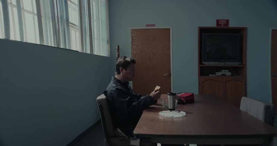 A man is sitting at a table in a room eating his lunch as he holds his sandwich with his hand on his cup