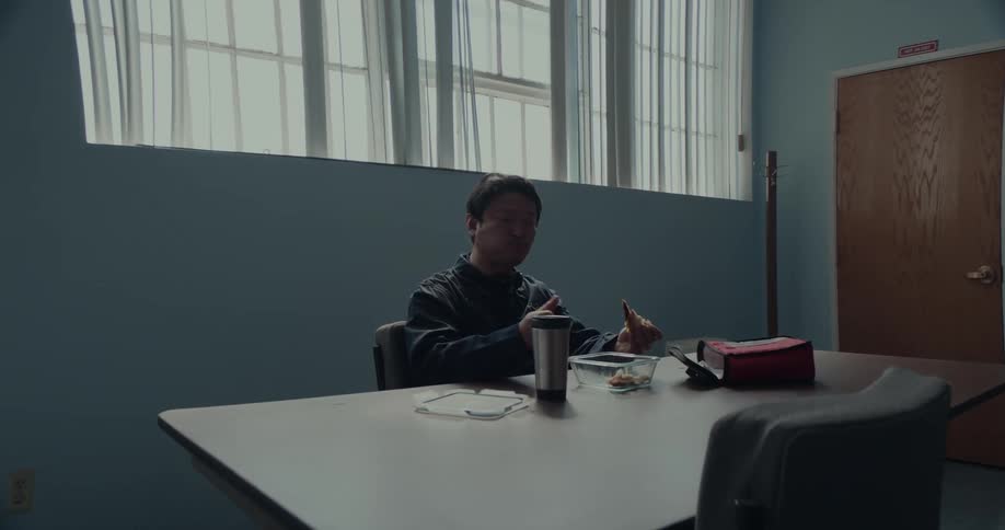 A man sits at a table under a row of windows in a room and holds a cup