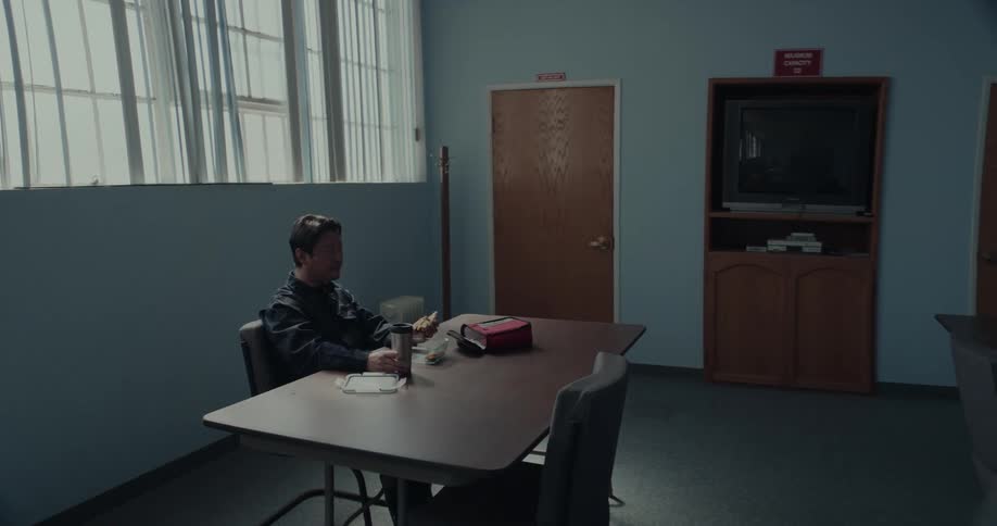 A man is sitting at a table in a room eating his lunch as he holds his sandwich as drinks from his cup