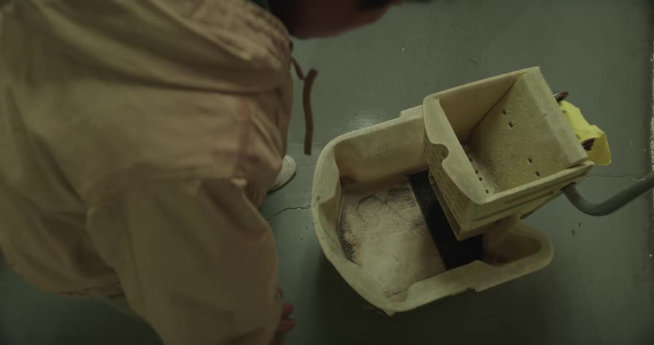 A man mops the floor then reaches in the mop bucket