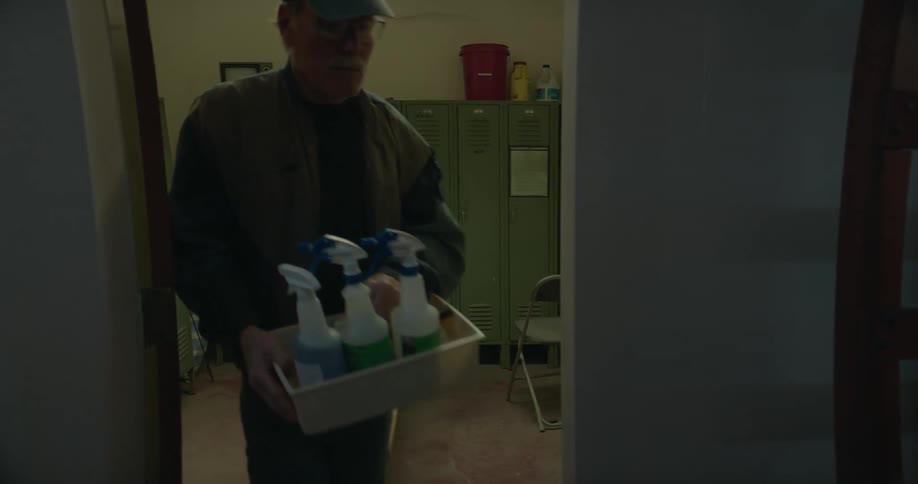 A janitor carries a tray of cleaning supplies out of a locker room as a man is sitting on a bench and another person walks out of the room