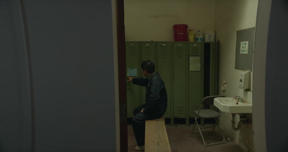 A janitor carries a tray of cleaning supplies out of a locker room as a man is sitting on a bench and he walks out of the room