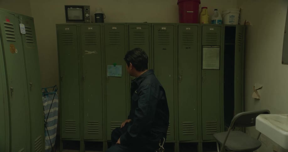 A janitor carries a tray of cleaning supplies out of a locker room and another person walks out of the room as a man sitting on a bench closes his locker and stands up and leaves