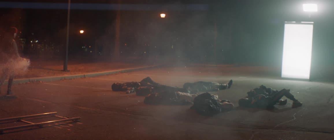 A prop man creates smoke around actors lying on the ground in a parking lot