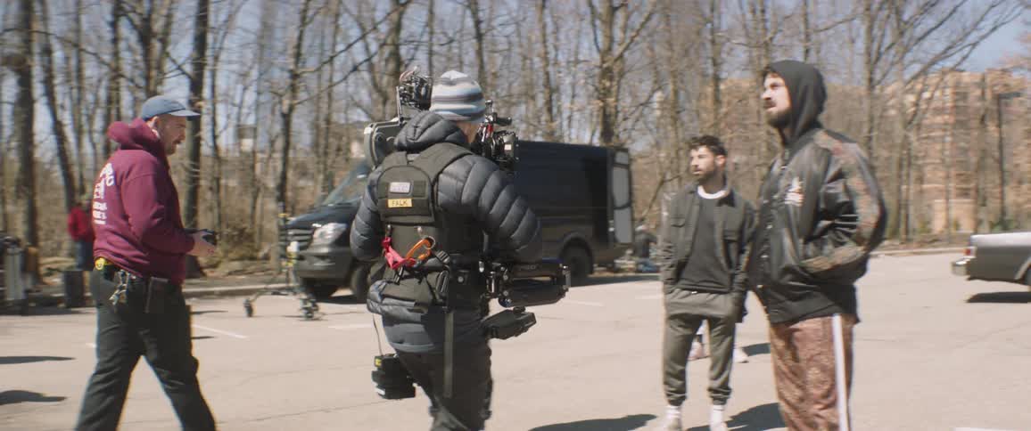 A camera man walks and films actors standing in a parking lot