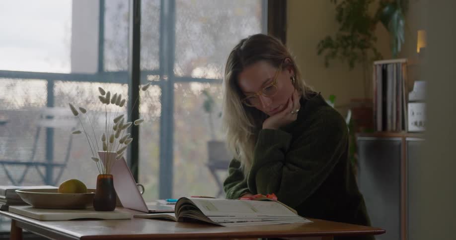 A woman sits at her desk reading a book while her laptop is open beside her