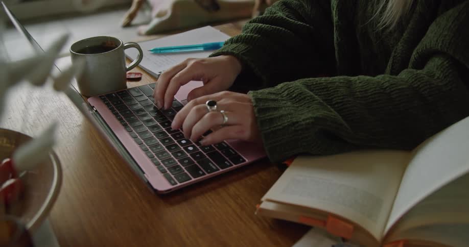 A woman looking through a book before turning to type on a laptop