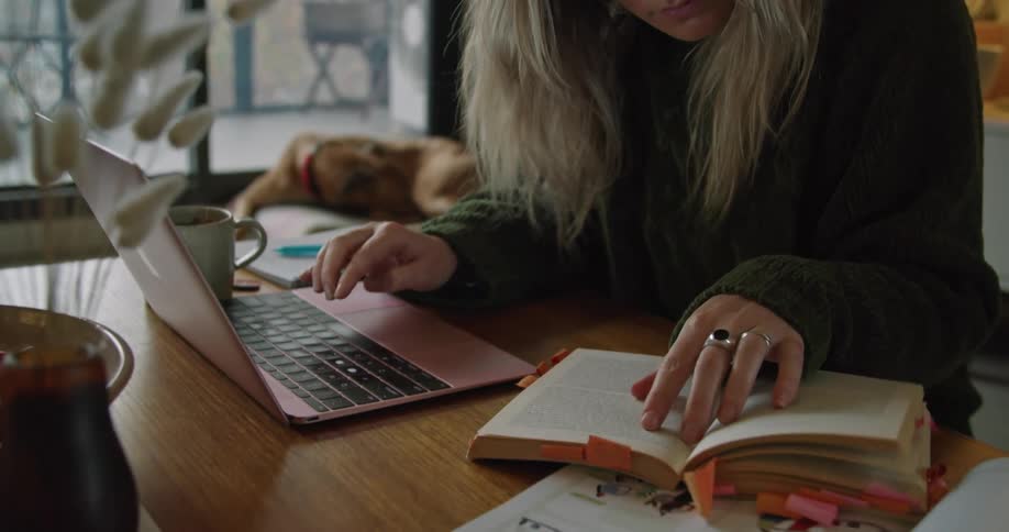 Woman sits at a desk working on a laptop and referring to a book