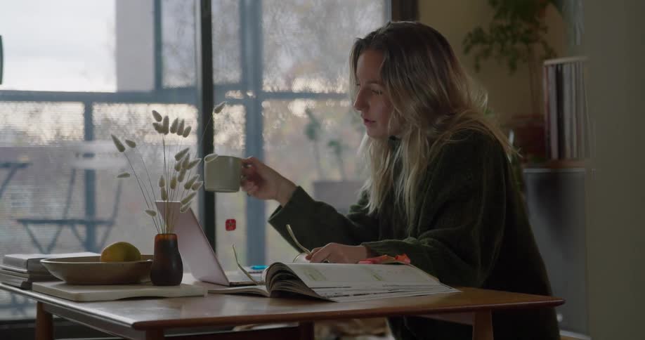 Woman drinks tea from a mug then puts on her glasses and looks at a notebook