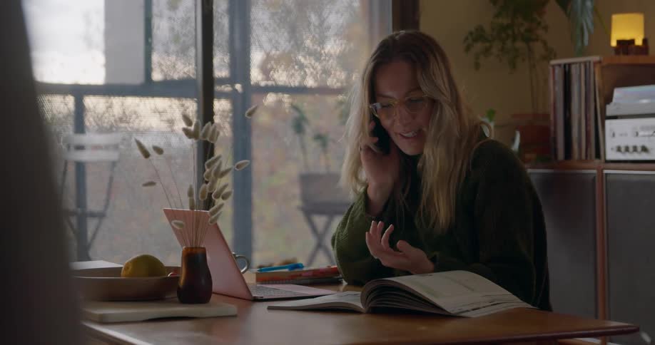 A woman talks on the phone while sitting at a desk and flipping through a magazine and looking at her laptop