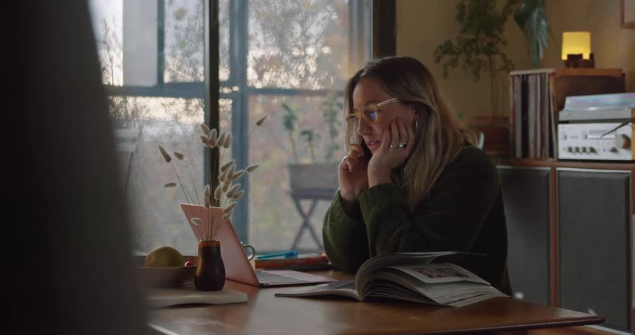 A white woman sits at a table in a living room while talking on the telephone