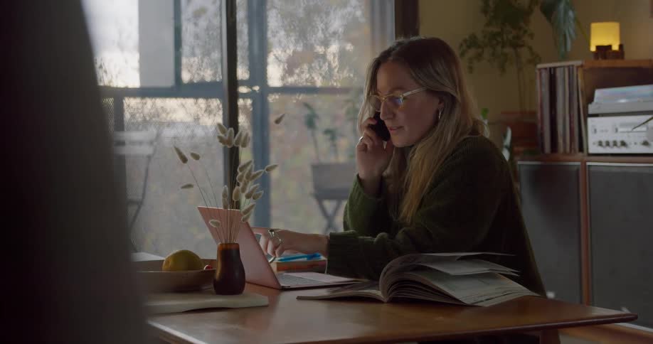 Woman sitting in living room works on laptop while talking on smartphone